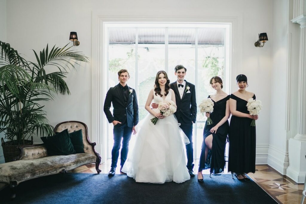 Couple's photo with their best man in black suit and bridesmaids in off-shoulder black gowns designed by Oleg Cassini