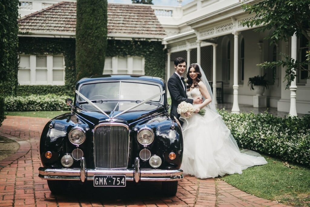 The couple posing next to their bridal car, a black 1955 classic Jaguar MKVII sedan